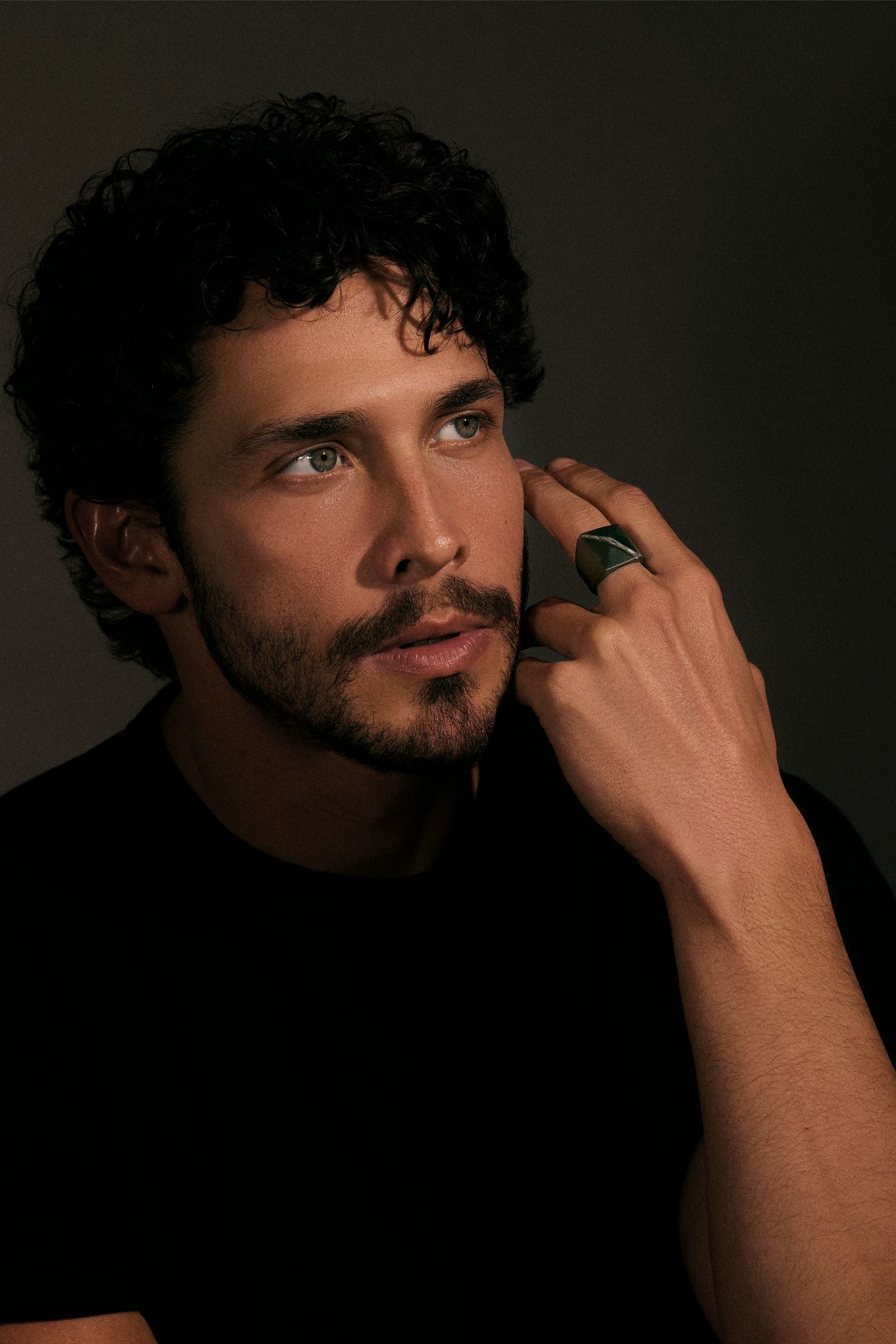 Man with a beard and curly hair wearing a black shirt, posing with hand near face against a dark background.