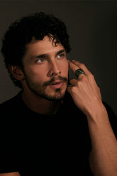 Man with a beard and curly hair wearing a black shirt, posing with hand near face against a dark background.
