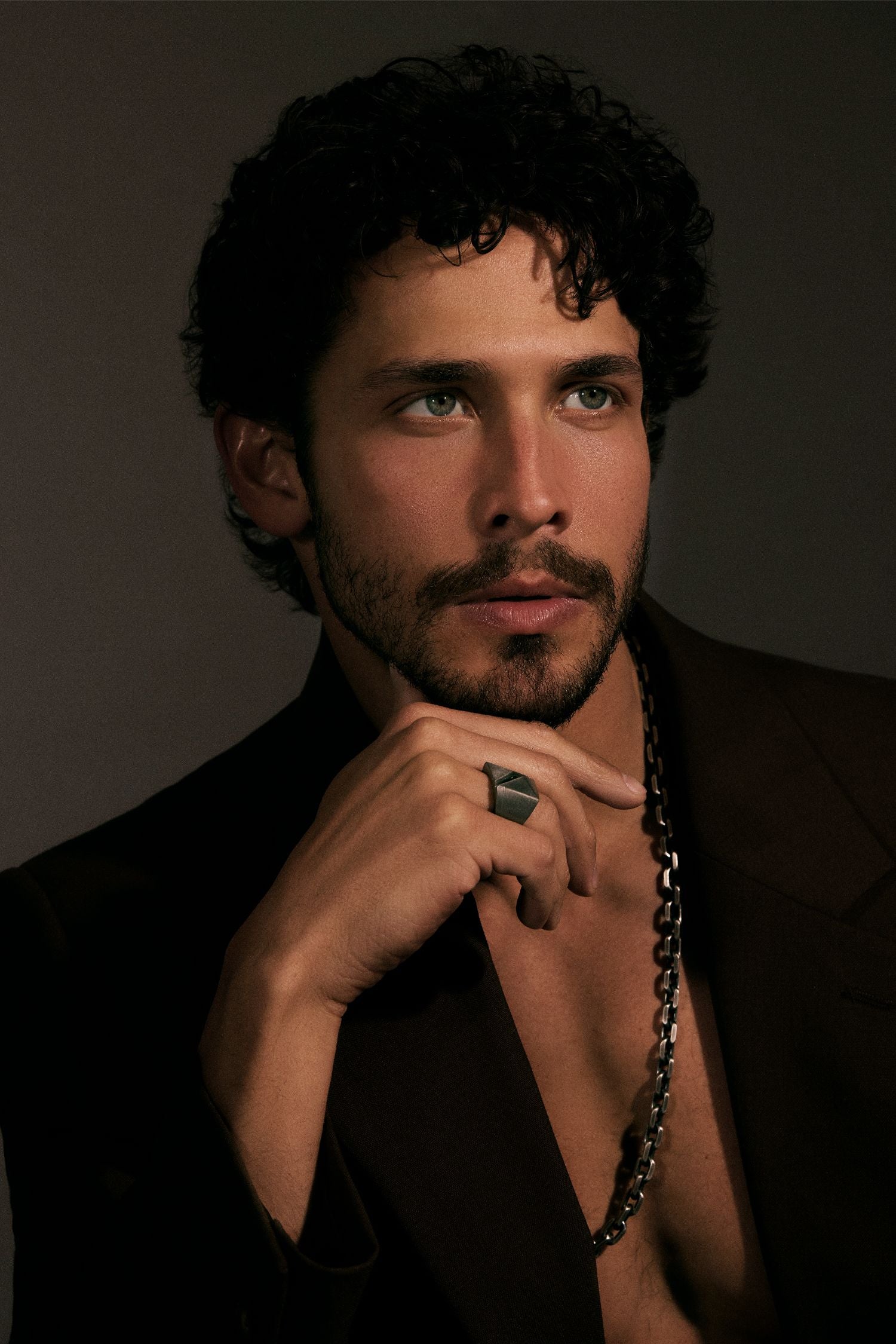Man with curly hair and beard wearing a dark suit against a dark background