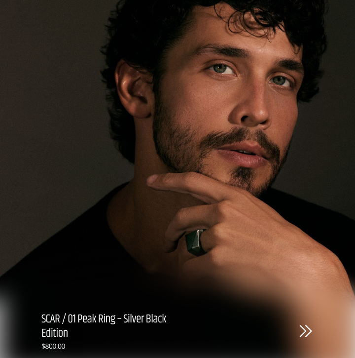 A close-up portrait of a man wearing a large silver black ring on his finger, with text overlay at the bottom reading 'SCAR / 01 Peak Ring - Silver Black Edition $800.00 Peak Ring